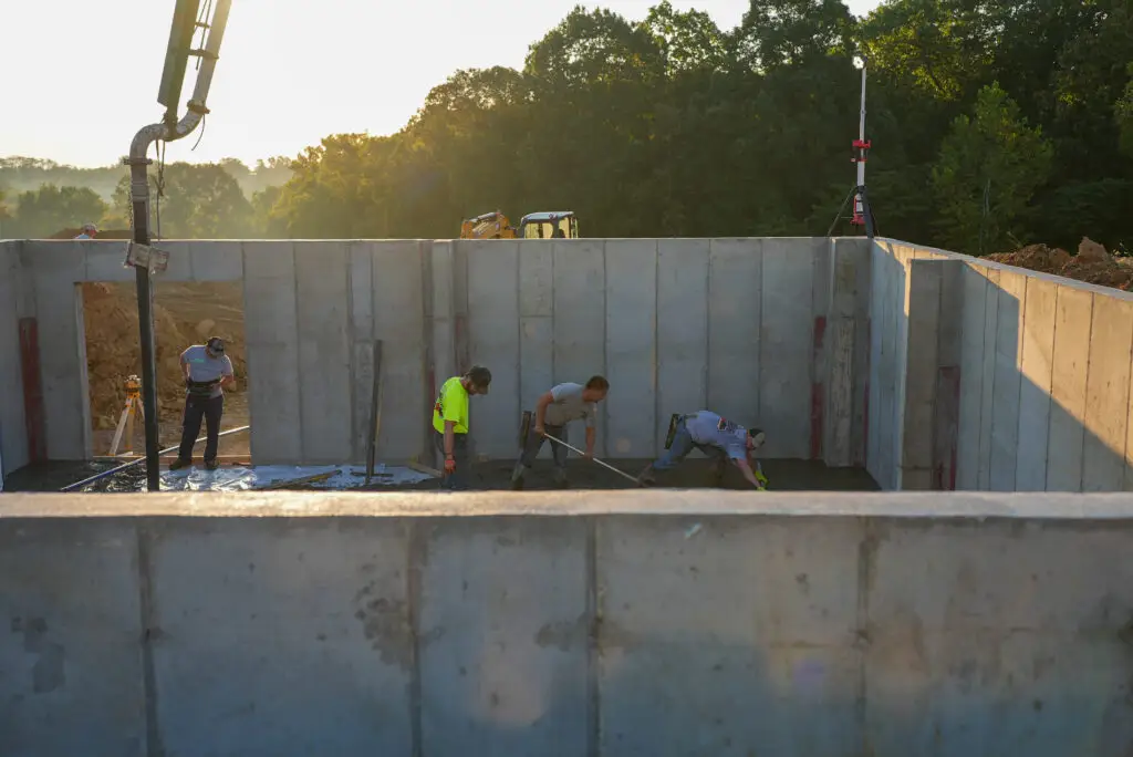 Construction workers in a concrete foundation pit, wearing hard hats, smoothing wet concrete with tools.