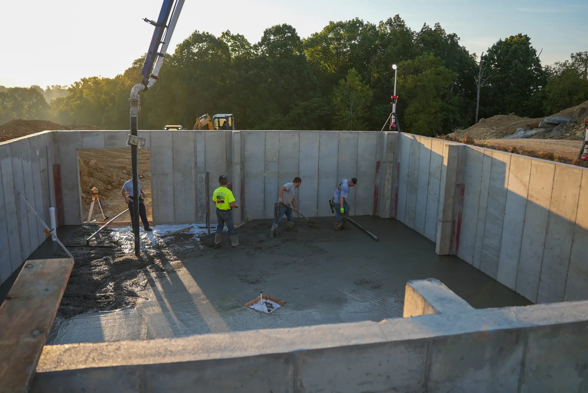Wide shot of Spencer's crew screeding and finishing a basement foundation slab pour at sunset, with the concrete pump hose visible at upper left