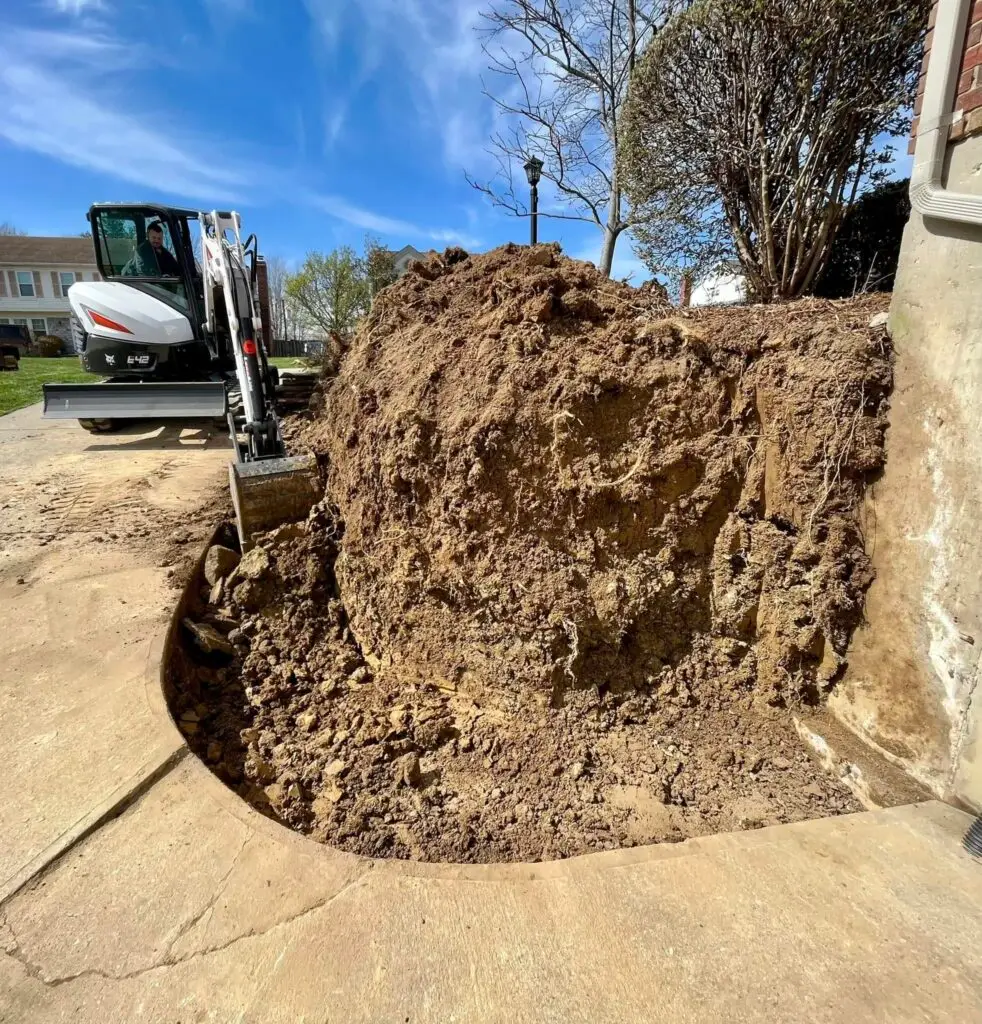 Bobcat compact excavator digging a large pit in a residential front yard alongside a concrete sidewalk