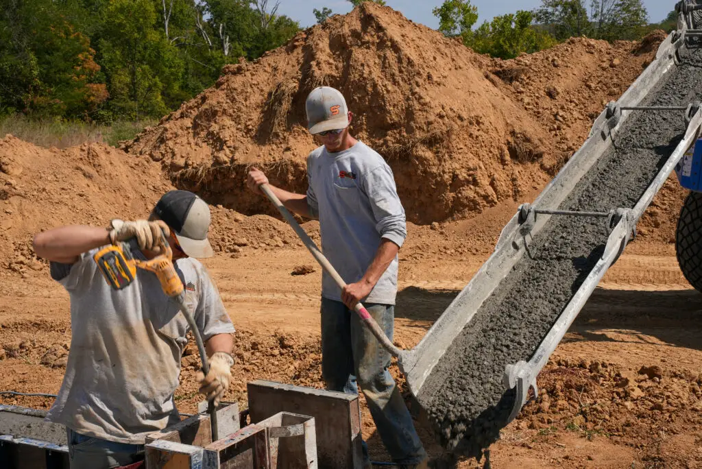 Two construction workers move dirt at a quarry site, with a large mound of soil and a gravel conveyor nearby.