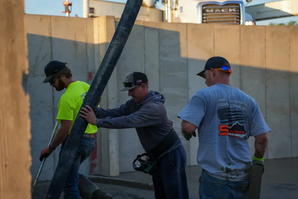 Workers in hard hats maneuver a large pipe between concrete forms at a construction site in warm evening light
