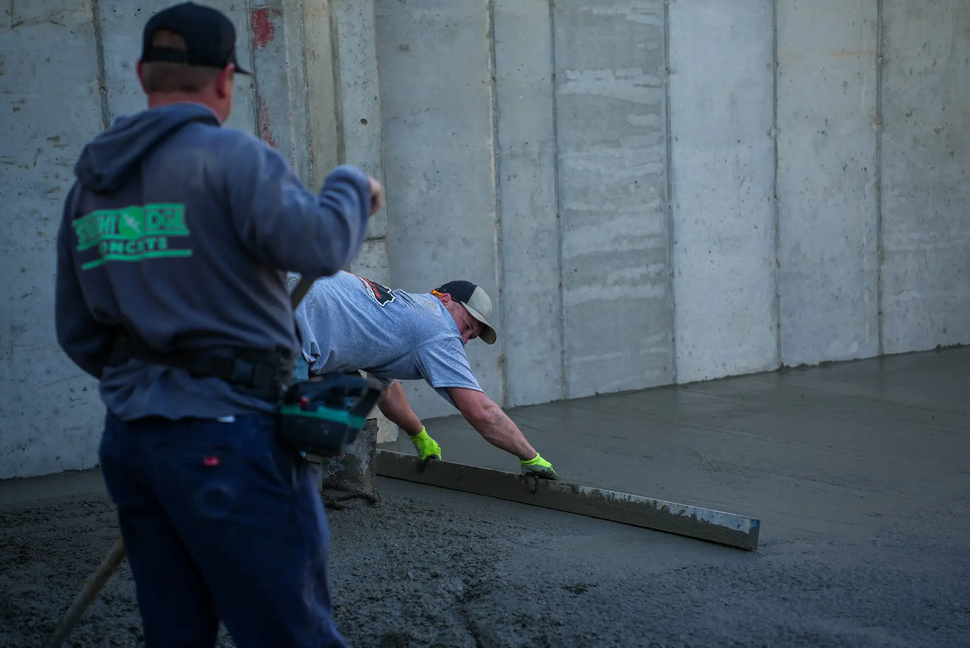 Two construction workers pour fresh concrete along a wooden form against a tall concrete wall, one standing behind the other.