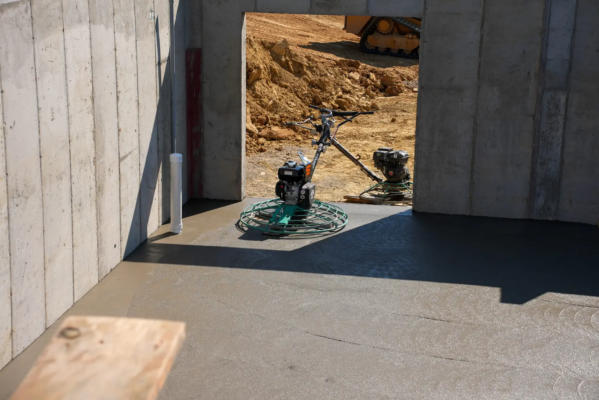 Spencer's walk-behind power trowel resting on a freshly finished basement concrete floor with a doorway to the dirt yard outside