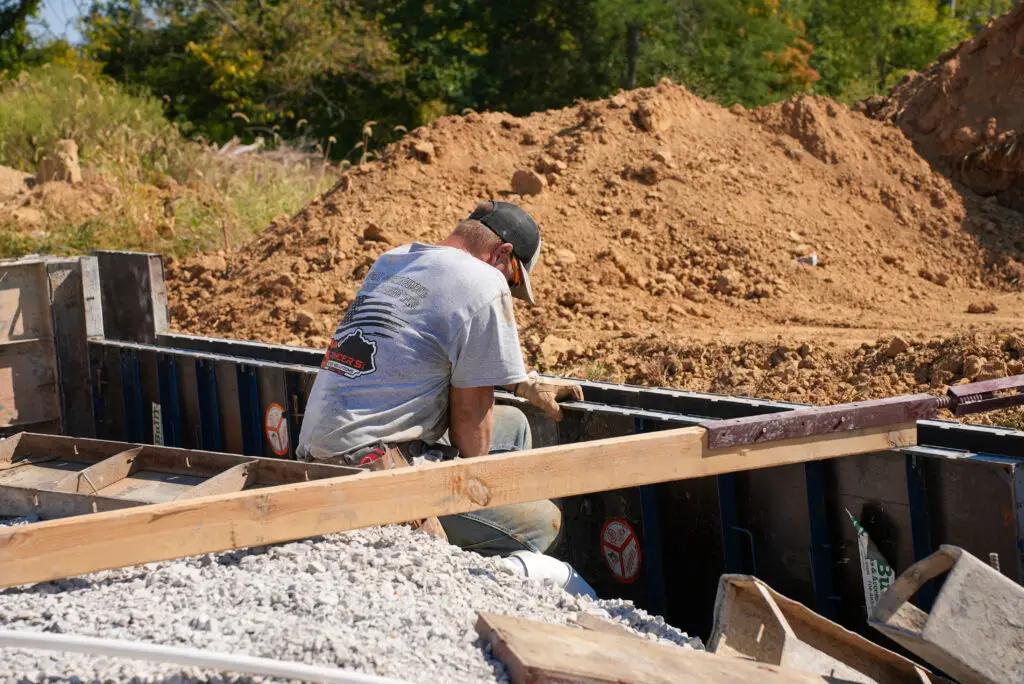 Construction worker kneeling and tying rebar in a wooden form at a dirt construction site with a large soil pile in the background