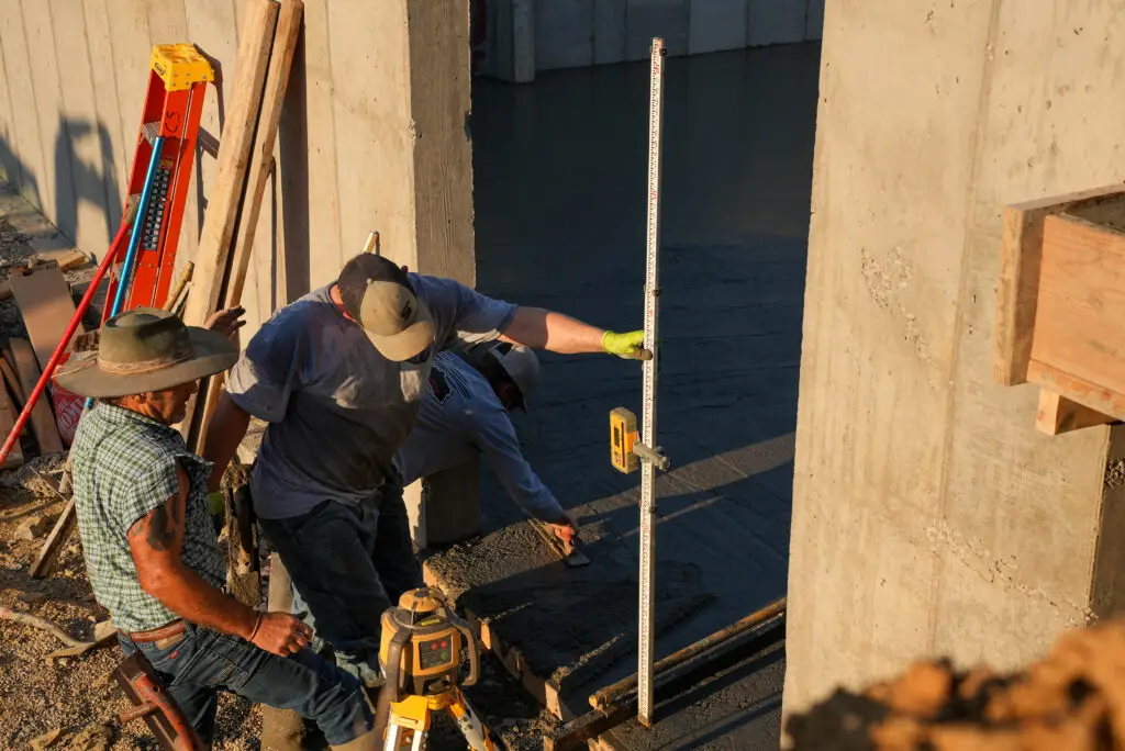 Two construction workers measure dimensions inside a concrete form with a tall vertical ruler on a sunny site, surrounded by tools and lumber.