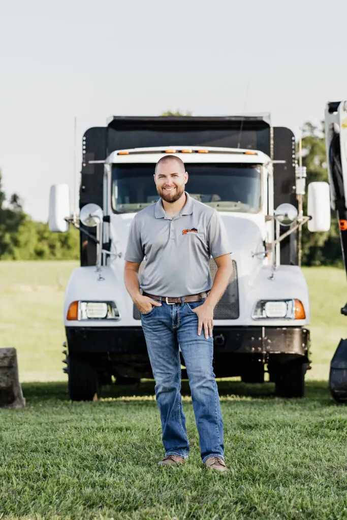 Man in a gray polo and jeans stands in front of a large white semi-truck outdoors on a grassy field, smiling at the camera.