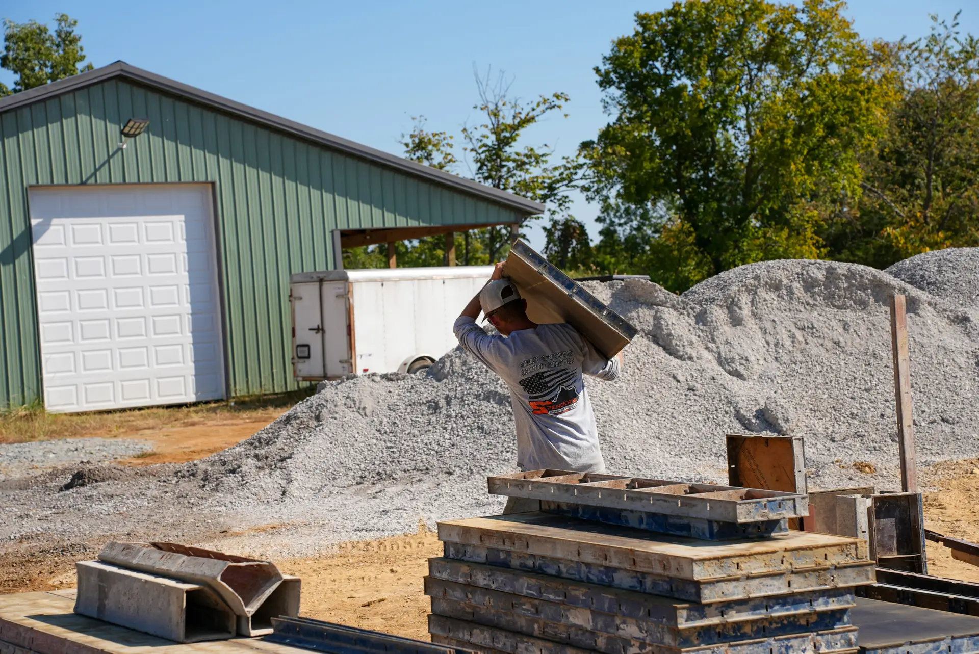 Construction worker lifts a large panel onto a stack of pallets at a gravel yard beside a green metal building and trailer, on a sunny day.