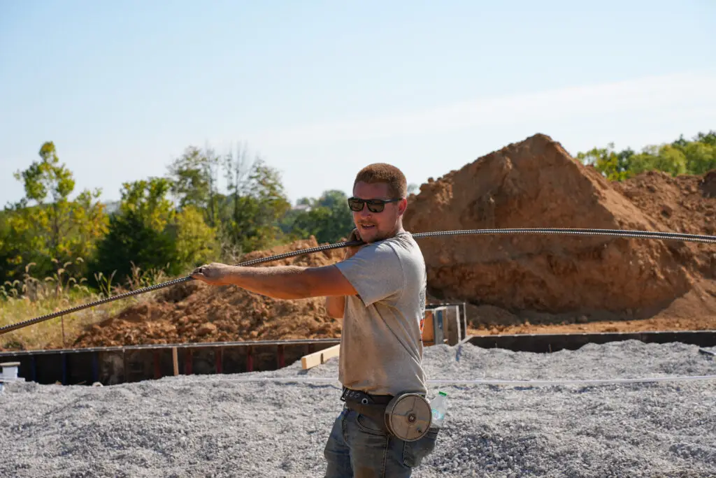 Construction worker in safety glasses pulls a steel cable across a gravel construction site with a dirt pile in the background.