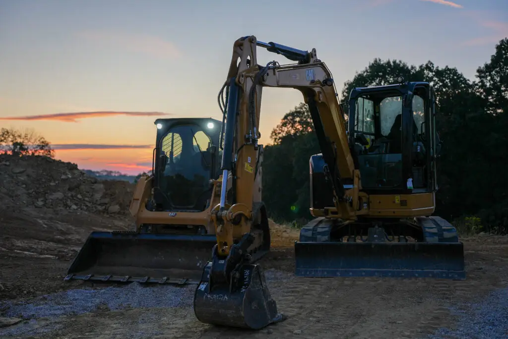 Two CAT excavators on a dirt construction site at sunset, with their buckets resting on the ground.