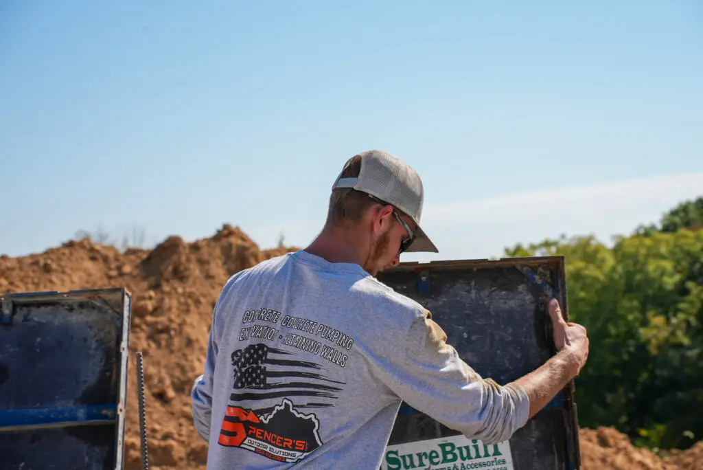 Construction worker in a beige cap leaning into a dumpster at a dirt construction site, loading materials.