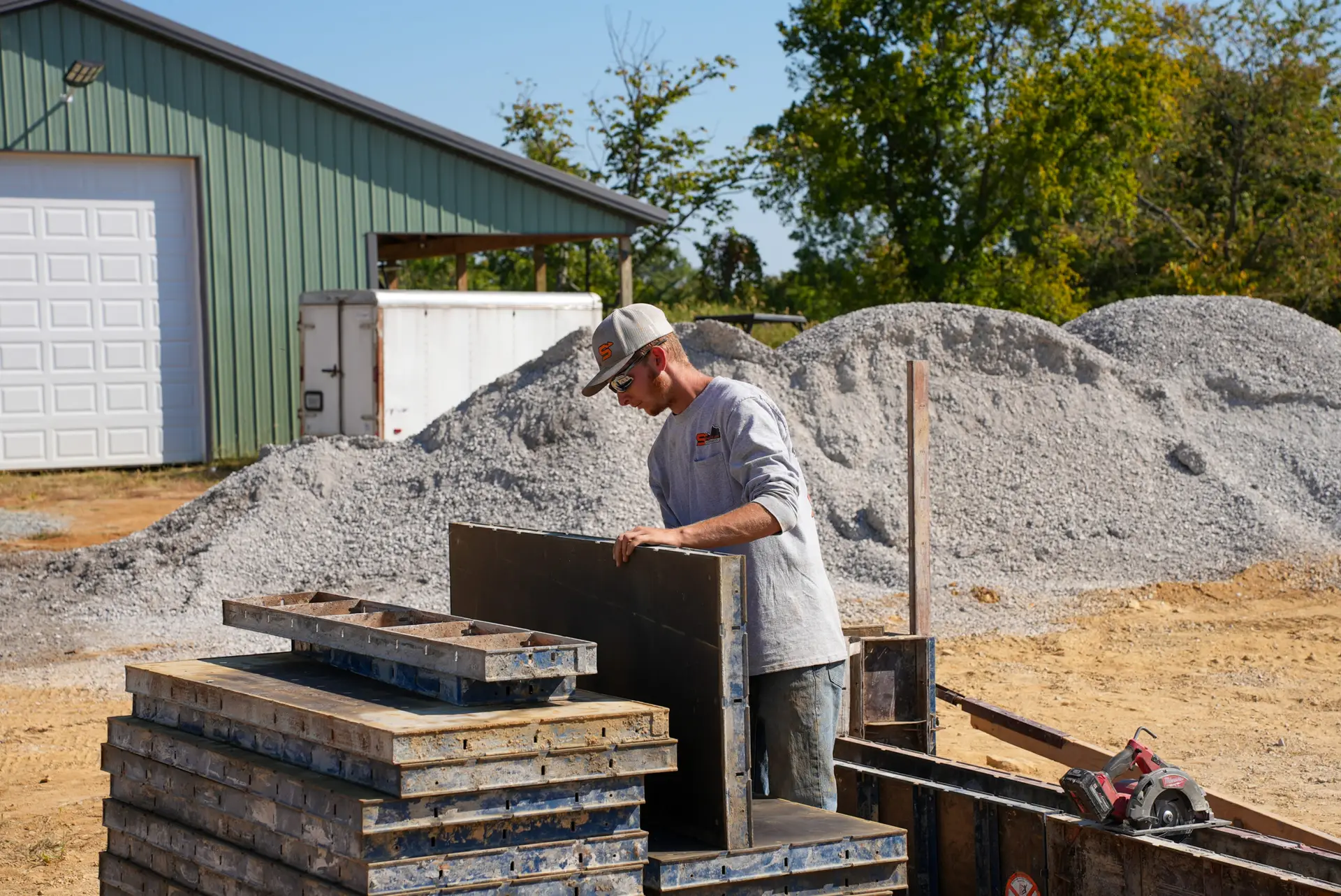 Construction worker in a gray shirt and cap assembles metal forms beside stacked pallets at an outdoor jobsite with gravel piles.